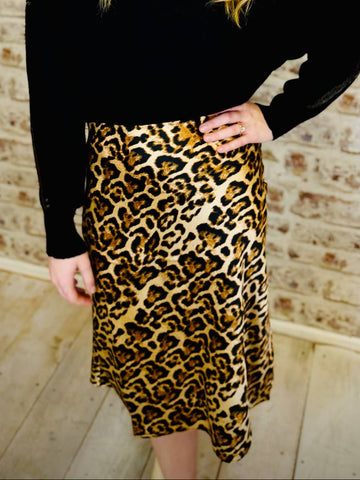 Person wearing a black top and leopard print skirt standing against a brick wall.